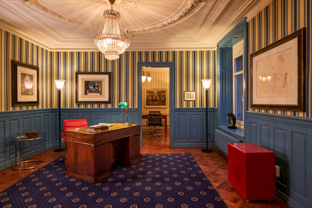 Salon Wohnzimmer mit elegantem Teppich, Holzschreibtisch und Kronleuchter in blau mit Streifentapete.